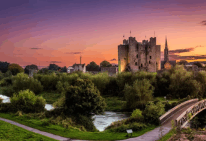 Medieval Irish castle near a river at sunset with surrounding greenery and bridge
